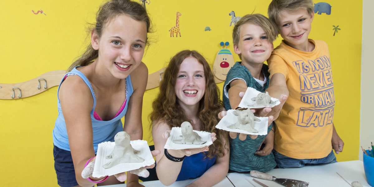 Vier Kinder halten in einem Klassenzimmer selbst gefertigte Tierskulpturen aus Ton hoch und lächeln in die Kamera. Kunstzubehör und bunte Zeichnungen schmücken den Tisch und die Wand hinter ihnen.