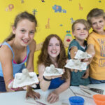 Vier Kinder halten in einem Klassenzimmer selbst gefertigte Tierskulpturen aus Ton hoch und lächeln in die Kamera. Kunstzubehör und bunte Zeichnungen schmücken den Tisch und die Wand hinter ihnen.