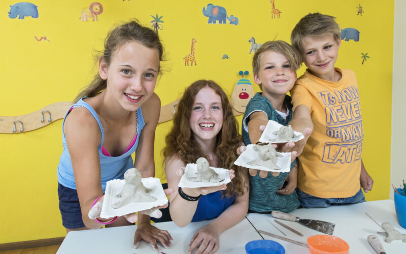 Vier Kinder halten in einem Klassenzimmer selbst gefertigte Tierskulpturen aus Ton hoch und lächeln in die Kamera. Kunstzubehör und bunte Zeichnungen schmücken den Tisch und die Wand hinter ihnen.