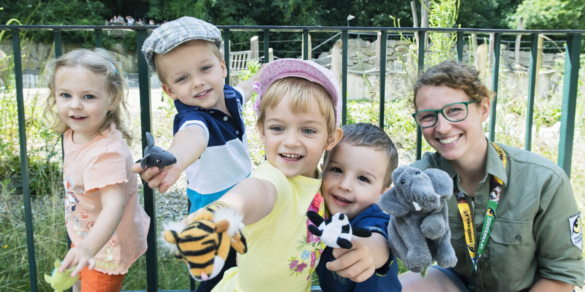 Vier kleine Kinder und ein Erwachsener posieren im Freien, lächeln und halten verschiedene Tierhandpuppen vor einem Metallzaun und Grünzeug hoch.