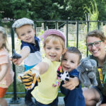 Vier kleine Kinder und ein Erwachsener posieren im Freien, lächeln und halten verschiedene Tierhandpuppen vor einem Metallzaun und Grünzeug hoch.