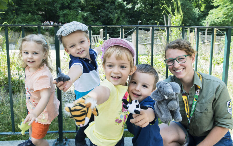 Vier kleine Kinder und ein Erwachsener posieren im Freien, lächeln und halten verschiedene Tierhandpuppen vor einem Metallzaun und Grünzeug hoch.