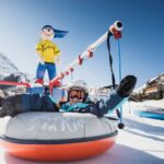 Eine Person in Winterkleidung fährt auf einem aufblasbaren Schneeschlauch und hält sich dabei an einem Zugseil fest. Im Hintergrund sind ein verschneiter Berg und eine Zeichentrickfigur zu sehen.
