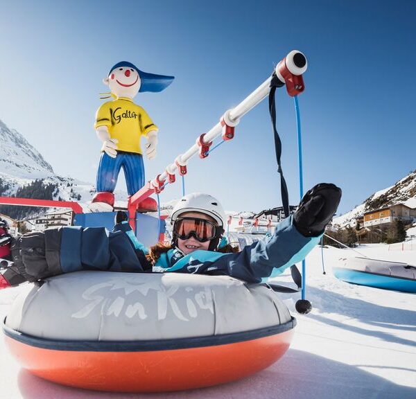 Eine Person in Winterkleidung fährt auf einem aufblasbaren Schneeschlauch und hält sich dabei an einem Zugseil fest. Im Hintergrund sind ein verschneiter Berg und eine Zeichentrickfigur zu sehen.