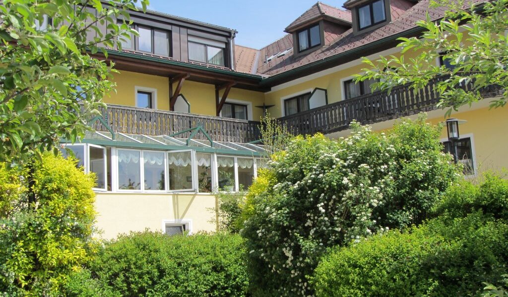 Ein großes gelbes Haus mit einem verglasten Wintergarten, dunklen Holzbalkonen und Dachgauben, umgeben von grünen Sträuchern und Bäumen.