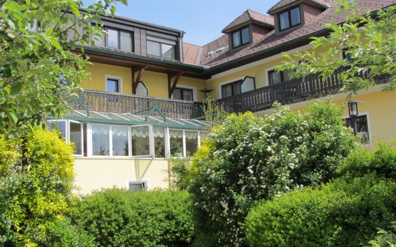 Ein großes gelbes Haus mit einem verglasten Wintergarten, dunklen Holzbalkonen und Dachgauben, umgeben von grünen Sträuchern und Bäumen.