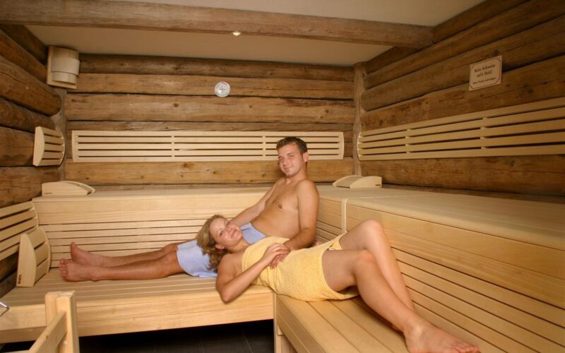 Zwei Personen entspannen auf Holzbänken in einer Blockbohlensauna, beide in Handtücher gehüllt.