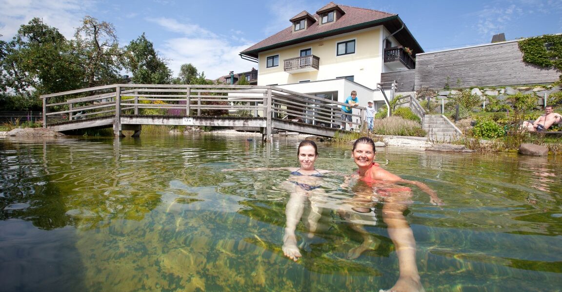 Zwei Personen schwimmen in einem klaren Teich vor einem Haus mit einer Holzbrücke und einem blauen Himmel im Hintergrund.