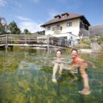 Zwei Personen schwimmen in einem klaren Teich vor einem Haus mit einer Holzbrücke und einem blauen Himmel im Hintergrund.