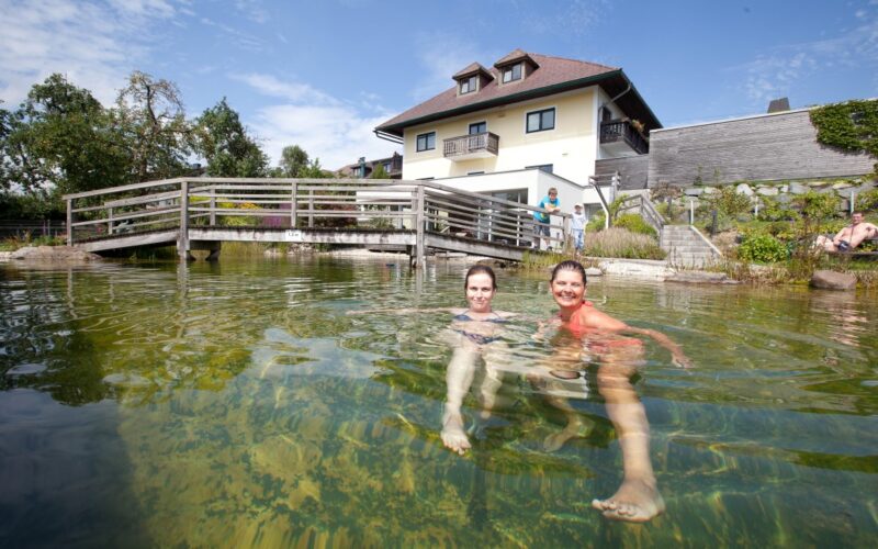 Zwei Personen schwimmen in einem klaren Teich vor einem Haus mit einer Holzbrücke und einem blauen Himmel im Hintergrund.