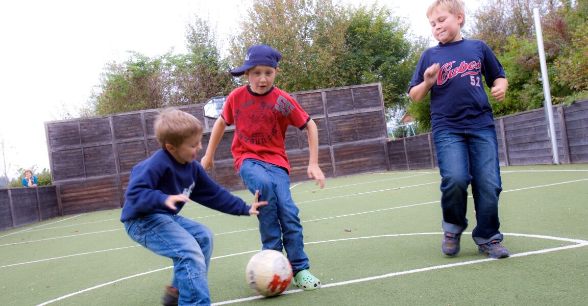 Drei Jungen spielen Fußball auf einem Kunstrasenplatz im Freien und konzentrieren sich darauf, einen weiß-roten Ball zu schießen.
