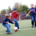 Drei Jungen spielen Fußball auf einem Kunstrasenplatz im Freien und konzentrieren sich darauf, einen weiß-roten Ball zu schießen.