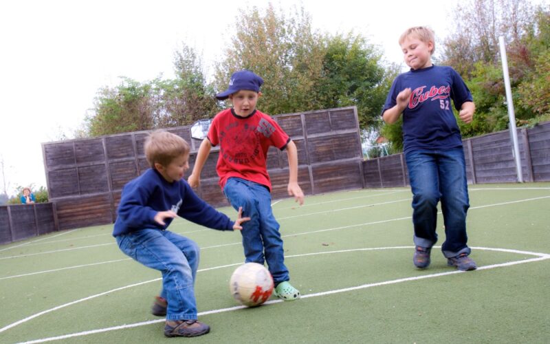 Drei Jungen spielen Fußball auf einem Kunstrasenplatz im Freien und konzentrieren sich darauf, einen weiß-roten Ball zu schießen.