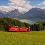 Ein roter Zug fährt durch ein grasbewachsenes Feld mit Bergen und einem See im Hintergrund unter einem teilweise bewölkten Himmel.