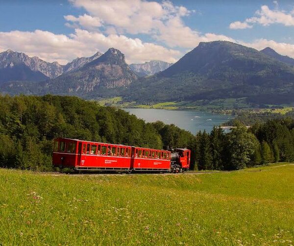 Ein roter Zug fährt durch ein grasbewachsenes Feld mit Bergen und einem See im Hintergrund unter einem teilweise bewölkten Himmel.