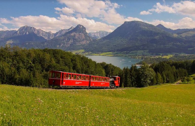 Ein roter Zug fährt durch ein grasbewachsenes Feld mit Bergen und einem See im Hintergrund unter einem teilweise bewölkten Himmel.