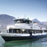 Ein weißes Fahrgastschiff mit dem Namen "Salzburg" fährt auf einem ruhigen See mit Bergen und klarem blauen Himmel im Hintergrund.