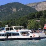 Ein weißes Fahrgastschiff mit einer österreichischen Flagge fährt auf einem türkisfarbenen See mit bewaldeten Hügeln und Häusern im Hintergrund.