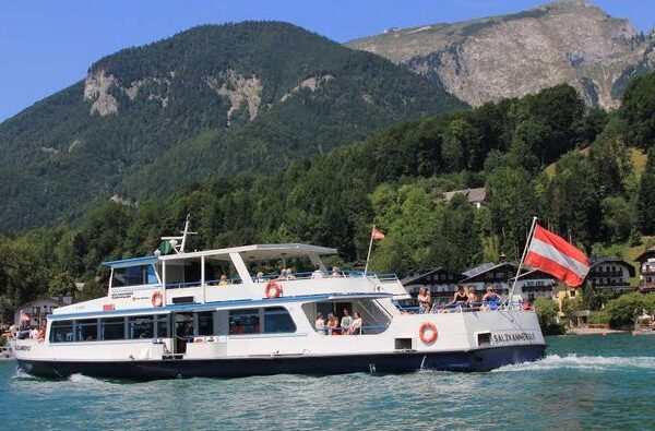 Ein weißes Fahrgastschiff mit einer österreichischen Flagge fährt auf einem türkisfarbenen See mit bewaldeten Hügeln und Häusern im Hintergrund.