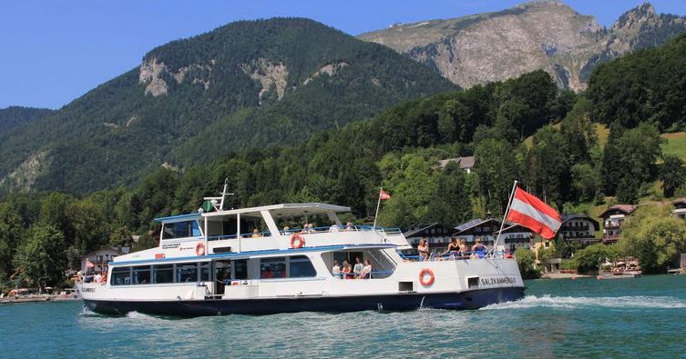 Ein weißes Fahrgastschiff mit einer österreichischen Flagge fährt auf einem türkisfarbenen See mit bewaldeten Hügeln und Häusern im Hintergrund.