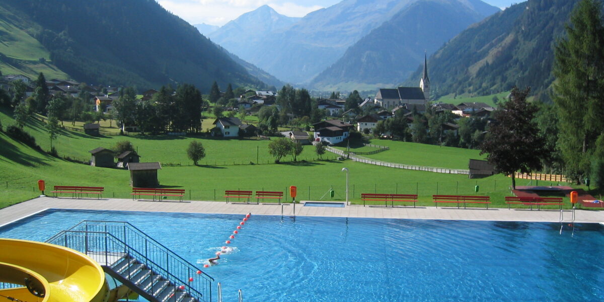 Außenschwimmbad mit gelber Rutsche, umgeben von Wiesen, Dorfhäusern und Bergen im Hintergrund unter einem blauen Himmel.