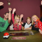 Vier Personen sitzen an einem Tisch mit einem "Team ZIRBE"-Schild und heben feierlich ihre Arme. Ein roter Buzzer, ein Spielbrett und Karten liegen auf dem Tisch.