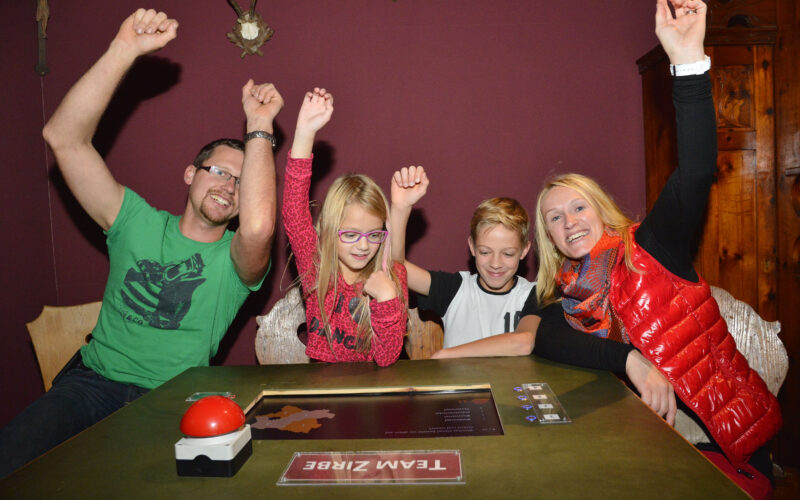 Vier Personen sitzen an einem Tisch mit einem "Team ZIRBE"-Schild und heben feierlich ihre Arme. Ein roter Buzzer, ein Spielbrett und Karten liegen auf dem Tisch.