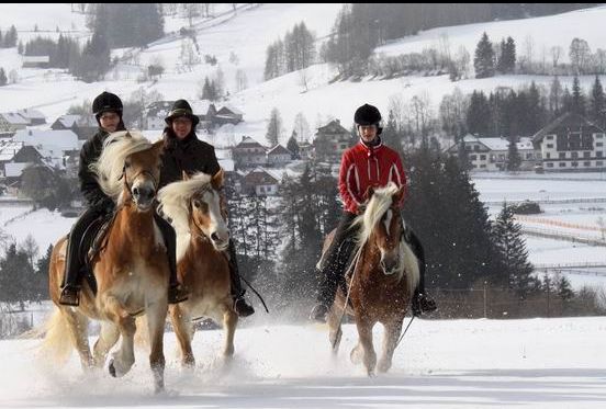 Drei Personen reiten auf Pferden durch Schnee in einer ländlichen, bergigen Gegend mit Häusern und Bäumen im Hintergrund.