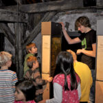 Eine Gruppe Kinder hört einem jungen Mann zu, der in einem rustikalen Holzgebäude an zwei vertikalen Anzeigetafeln etwas erklärt.