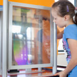 Ein junges Mädchen in einem blauen Hemd betrachtet ein wissenschaftliches Exponat durch eine transparente Scheibe, mit orangefarbenen Silhouetten im Hintergrund.