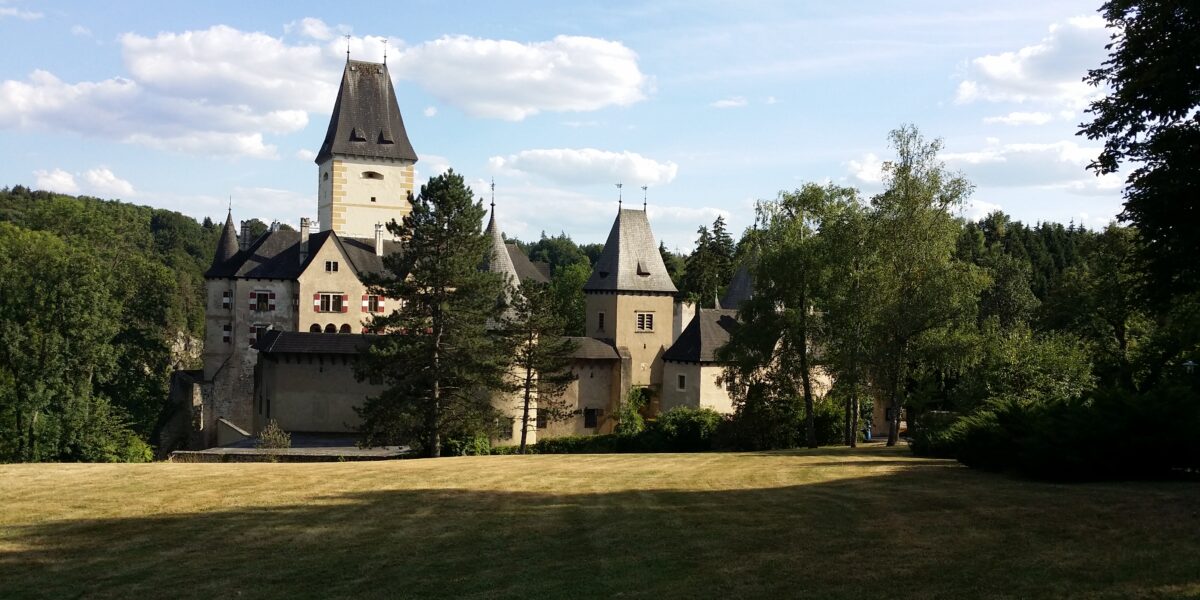 Eine historische Steinburg mit hohen Türmen und steilen Dächern steht umgeben von Bäumen und einer großen Wiese unter einem teilweise bewölkten Himmel.