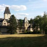 Eine historische Steinburg mit hohen Türmen und steilen Dächern steht umgeben von Bäumen und einer großen Wiese unter einem teilweise bewölkten Himmel.