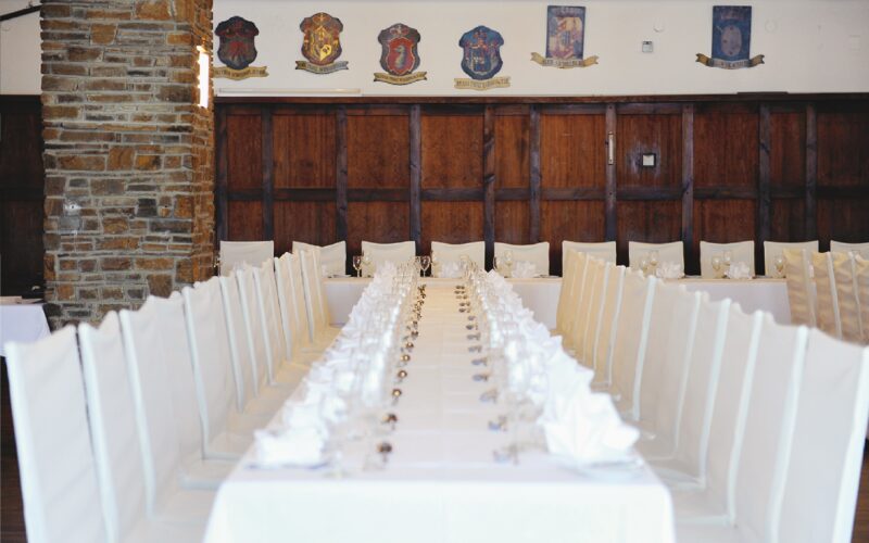 Ein langer Esstisch mit weißen Stühlen und Tischtüchern, der für eine formelle Veranstaltung gedeckt ist, mit Holzvertäfelung und dekorativen Wappen an der Wand im Hintergrund.