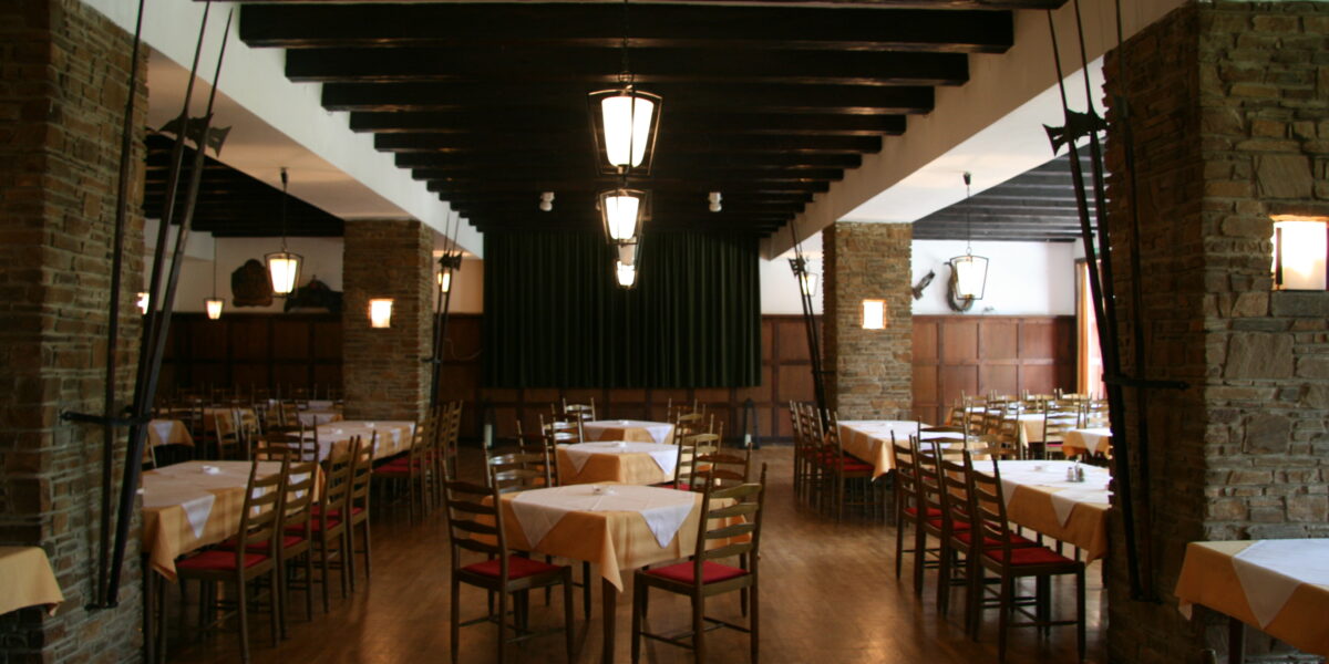 Geräumiger Speisesaal des Restaurants mit Holzböden, Steinsäulen, leeren Tischen mit gelben Tischtüchern und ordentlich aufgestellten Holzstühlen. Deckenbalken und Hängelampen sind sichtbar.