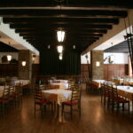 Geräumiger Speisesaal des Restaurants mit Holzböden, Steinsäulen, leeren Tischen mit gelben Tischtüchern und ordentlich aufgestellten Holzstühlen. Deckenbalken und Hängelampen sind sichtbar.