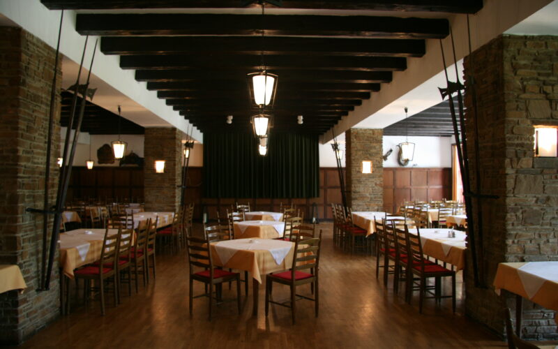 Geräumiger Speisesaal des Restaurants mit Holzböden, Steinsäulen, leeren Tischen mit gelben Tischtüchern und ordentlich aufgestellten Holzstühlen. Deckenbalken und Hängelampen sind sichtbar.