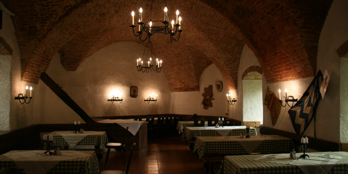 Schummerig beleuchtetes Restaurant mit gewölbter Ziegeldecke, Kronleuchtern und Tischen mit karierten Tischdecken, die im ganzen Raum verteilt sind.