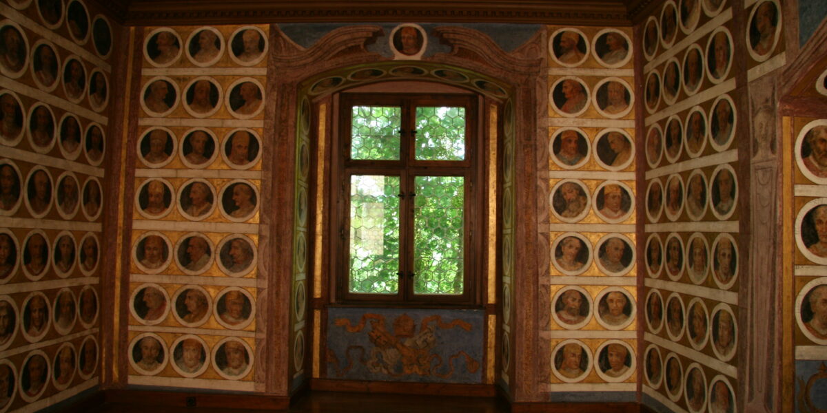Ein reich dekorierter Raum, dessen Wände mit kreisförmigen Porträtgemälden bedeckt sind, die ein zentrales Fenster umgeben, das grünes Laub nach draußen zeigt.