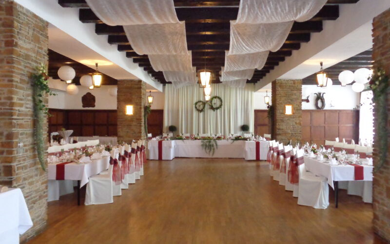 Ein Bankettsaal mit geschmückten Tischen und Stühlen, weißen Deckenvorhängen, Holzböden, Backsteinsäulen und einem mit Grünzeug und Kränzen geschmückten Haupttisch am Ende.