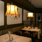 Ein gemütliches Restaurant mit gemusterten Bänken, weißen Tischtüchern, gefalteten Servietten, Blumenvasen und warmen Hängelampen an der Decke.
