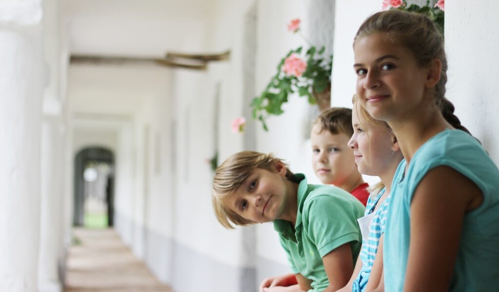 Vier Kinder sitzen auf einer Bank in einem hellen Korridor mit weißen Wänden und rosa Blumen und schauen in die Kamera.