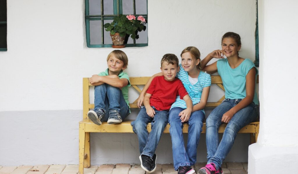 Vier Kinder sitzen auf einer Holzbank vor einem weißen Gebäude, mit einem Fenster und einer Topfpflanze im Hintergrund.