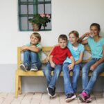 Vier Kinder sitzen auf einer Holzbank vor einem weißen Gebäude, mit einem Fenster und einer Topfpflanze im Hintergrund.