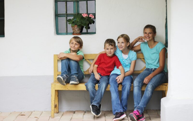 Vier Kinder sitzen auf einer Holzbank vor einem weißen Gebäude, mit einem Fenster und einer Topfpflanze im Hintergrund.