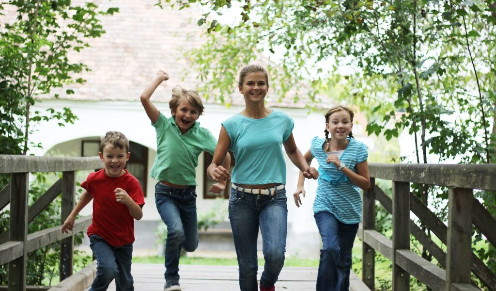 Vier Kinder in Freizeitkleidung laufen gemeinsam im Freien über eine Holzbrücke, umgeben von grünen Bäumen.