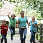 Vier Kinder in Freizeitkleidung laufen gemeinsam im Freien über eine Holzbrücke, umgeben von grünen Bäumen.