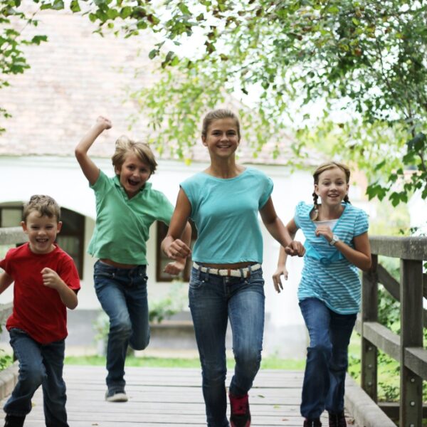 Vier Kinder in Freizeitkleidung laufen gemeinsam im Freien über eine Holzbrücke, umgeben von grünen Bäumen.