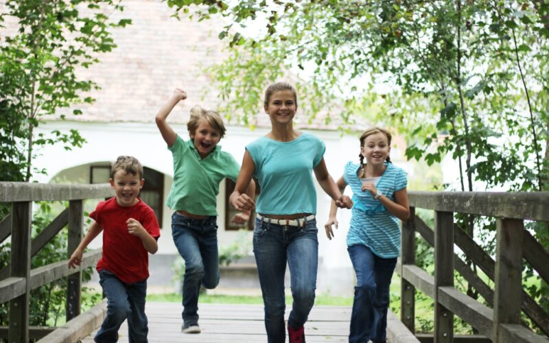 Vier Kinder in Freizeitkleidung laufen gemeinsam im Freien über eine Holzbrücke, umgeben von grünen Bäumen.