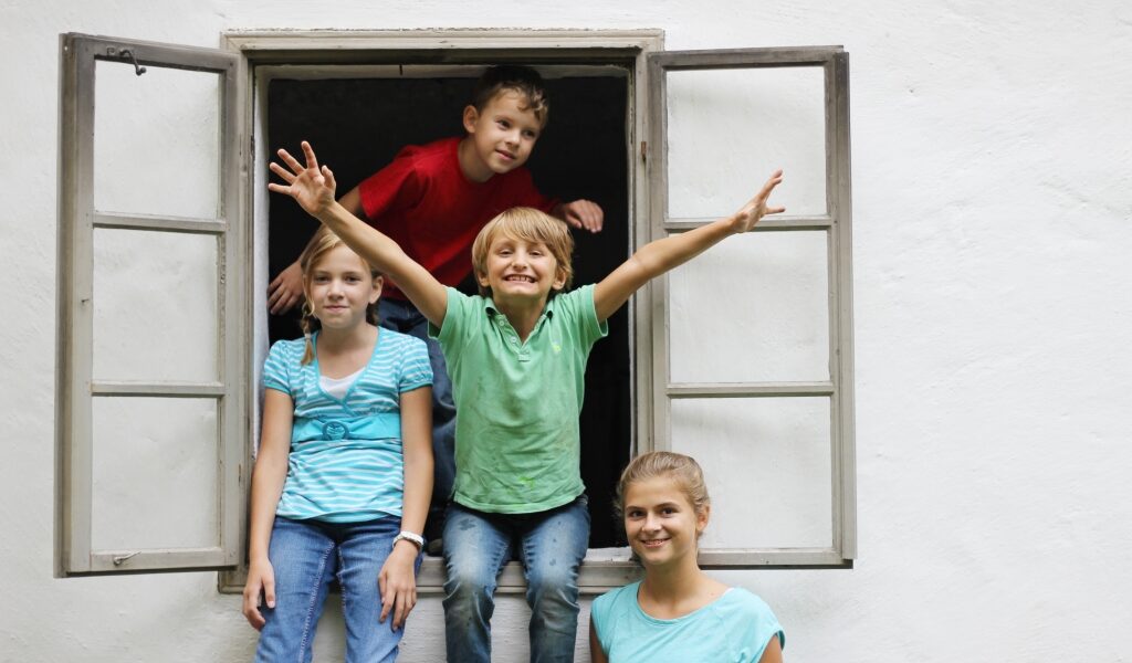 Vier Kinder posieren und lächeln vor einem Fenster mit weißer Wand, zwei sitzen auf der Fensterbank und zwei stehen dahinter, wobei ein Kind begeistert die Arme hebt.