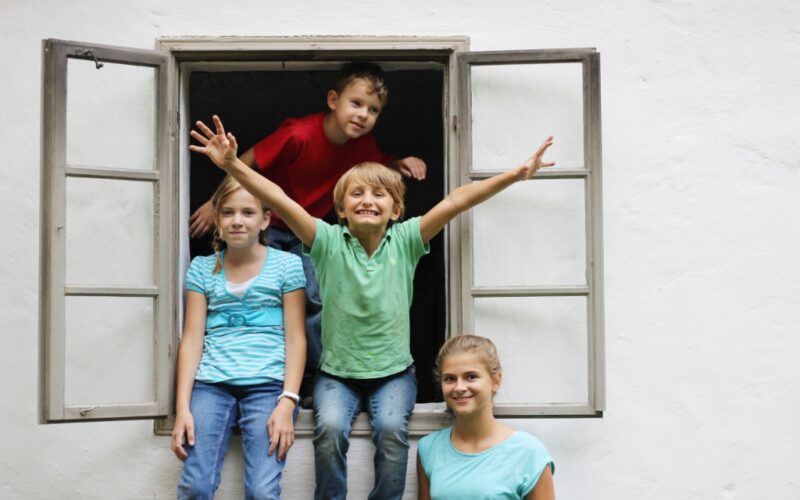 Vier Kinder posieren und lächeln vor einem Fenster mit weißer Wand, zwei sitzen auf der Fensterbank und zwei stehen dahinter, wobei ein Kind begeistert die Arme hebt.